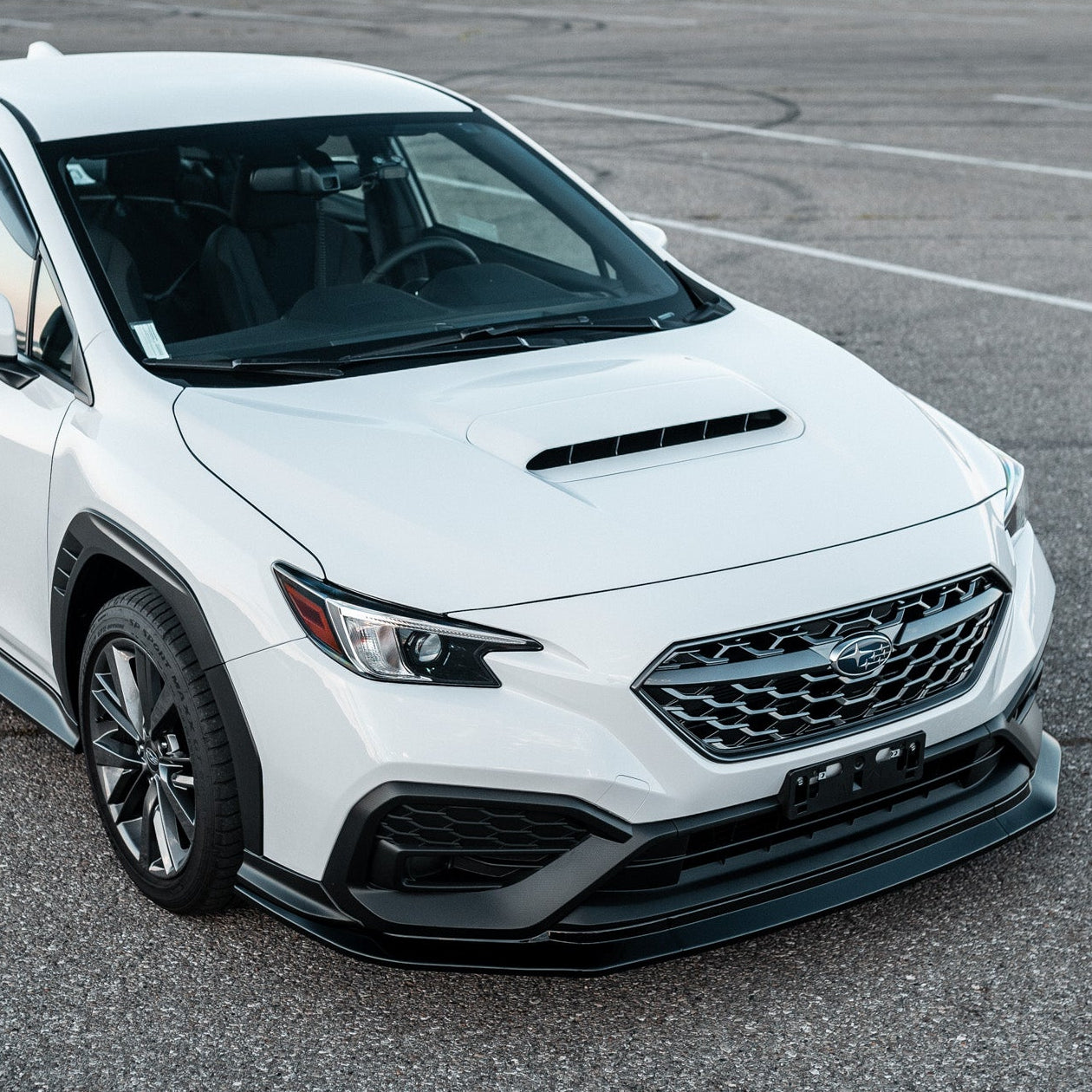 A white Subaru vehicle parked on an asphalt surface with a body kit, prominent front grille and logo, and visible license plate.